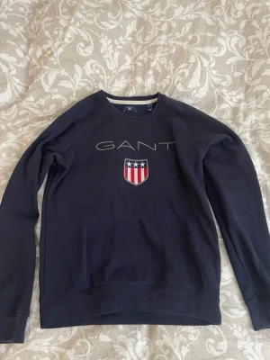 Mörkblå sweatshirt från GANT - Säljer en mörkblå sweatshirt från GANT med klassisk logga och broderad sköld med stjärnor och ränder på bröstet. Tröjan har rund halsringning och långa ärmar. Perfekt för en stilren och avslappnad look.