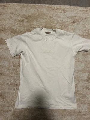 Vit Burberry t-shirt med broderi - Vit t-shirt från Burberry med broderad logga framtill och stor broderad Burberry-text på ryggen. Klassisk passform och rund halsringning. Tillverkad i mjuk bomull, perfekt för en stilren och enkel look.