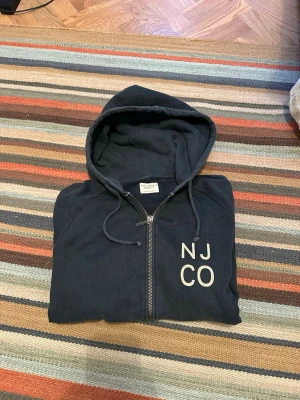 Mörkblå hoodie från Nudie Jeans n Company - Snygg hoodie från Nudie Jeans n Company i svart/grå färg. Lite vintage inspirerad modell med klassisk huva, kängurufickor och NJCO på bröstet.