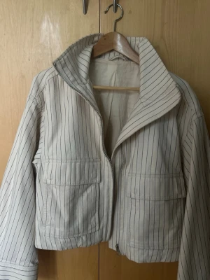 Vit jacka med svarta ränder - Snygg vit jacka med tunna svarta vertikala ränder och dragkedja framtill. Jackan har två stora fickor på framsidan och en hög krage. Materialet känns som bomull och modellen är boxig och croppad, perfekt för en trendig look.