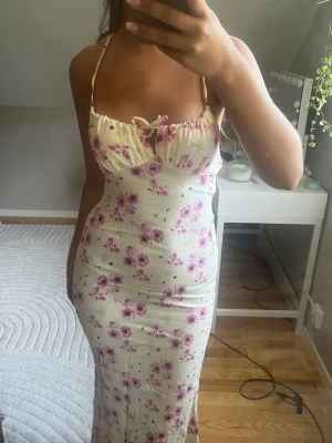 Lång blommig klänning med snörning - Supersöt långklänning i ljus färg med rosa blommor. Klänningen har smala axelband, snörning fram och öppen rygg med korsade band. Figurnära passform och volangdetalj vid bysten. Perfekt för sommarens alla tillfällen!