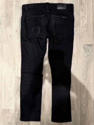 Nudie Jeans Lean Dean Svarta Selvage W29/L28 - Slim Tapered Fit - Nudie Jeans Lean Dean ”Dry Black Selvage” svarta selvage-jeans med slim tapered-passform, normalhög midja och avsmalnande ben, storlek W29/L28, midja 39,5 cm, ytterbenslängd 91 cm, benöppning 15,5 cm, modellen är 185cm & väger 65kg, i fint skick.