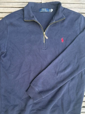 Marinblå stickad tröja Polo Ralph Lauren - Snygg marinblå stickad tröja från Polo Ralph Lauren med halv dragkedja och klassisk röd logga på bröstet. Tröjan har lång ärm, hög krage och ribbade muddar. Perfekt till hösten för lager på lager och enkel att matcha till olika outfits.