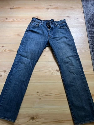 Levi's 501 raka blå jeans W32 L32 - Klassiska Levi's 501 jeans i blå denim med rak passform och knappgylf. Jeansen har fem fickor och den ikoniska läderpatchen bak i midjan. Perfekta för en avslappnad och tidlös stil.