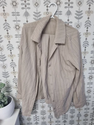 Beige randig skjorta med knappar - Säljer en beige skjorta med diskreta ränder och knappar framtill. Skjortan har lång ärm, klassisk krage och är tillverkad i ett mjukt tyg som känns luftigt och bekvämt. Perfekt för dig som gillar en avslappnad men stilren look.
