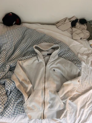 Vit hoodie från Polo Ralph Lauren - Snygg vit hoodie från Polo Ralph Lauren med dragkedja framtill och klassisk huva. Tröjan har två fickor fram och den ikoniska lilla loggan broderad på bröstet. Perfekt för en chill och avslappnad stil. Tillverkad i mjukt bomullsmaterial. De finns två små hål på båda armbågar och lite småfläckar