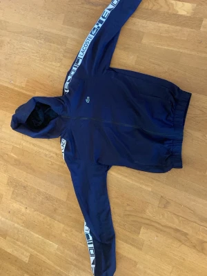 Marinblå vindjacka från Lacoste - Snygg marinblå vindjacka från Lacoste är storlek L men är som small elle medium. med huva och dragkedja. Jackan har vita detaljer och logga längs ärmarna samt den klassiska krokodilloggan på bröstet. Tillverkad i lätt och vindtätt material, perfekt för blåsiga dagar.