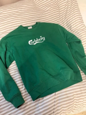 Grön crewneck Carlsberg sweatshirt - Snygg grön crewneck sweatshirt med Carlsberg-tryck på bröstet och 'SUMMERON CREW' på ryggen. Klassisk passform med ribbade muddar vid ärmar och nederkant. Perfekt för dig som gillar streetwear och vill sticka ut lite extra.