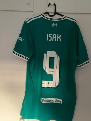  Liverpool matchtröja Isak 9 - Säljer en grön Liverpool fotbollströja med vita detaljer och tryck. Tröjan har korta ärmar, klubbmärke och sponsorloggor på bröstet samt Champions League-märke på ärmen. Namnet ISAK och nummer 9 är tryckt på ryggen. Perfekt för dig som älskar fotboll och vill sticka ut på planen.