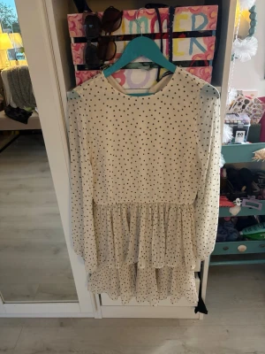 Vit prickig volangklänning - Supersöt vit kortklänning med svarta prickar och volangdetaljer. Klänningen har långärmade ärmar och rund halsringning. Tillverkad i ett lätt och luftigt material, perfekt för varmare dagar.