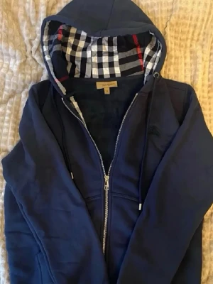 Marinblå hoodie med rutigt foder - Snygg marinblå hoodie med dragkedja och huva. Insidan av huvan har ett klassiskt rutigt mönster i vitt, svart och rött. Hoodien har dragsko i huvan och fickor framtill. Perfekt för en chill och stilren look.