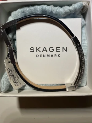 Svart läderarmband från Skagen - Stilrent armband från Skagen i svart läder med detaljer i rostfritt stål. Armbandet har en elegant metallplatta med diskret textur och Skagen-logga. Snyggt och minimalistiskt designat, perfekt för dig som gillar en clean look.