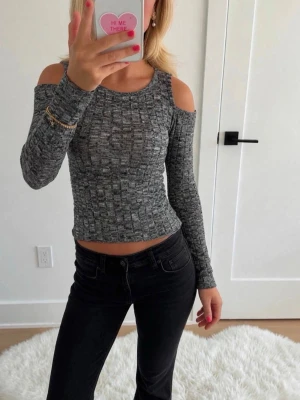 Grå ribbad topp med cut out-axlar - Trendig grå långärmad topp med ribbad struktur och snygga cut out-detaljer vid axlarna. Figurnära passform som framhäver siluetten och ger en modern look. Perfekt att styla med både jeans och kjol för en cool vibe.