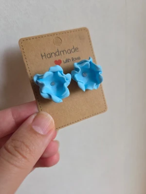 Blå blommiga örhängen i lera - Handgjorda örhängen i form av blå blommor med vågiga kanter. Tillverkade av mig i lera och har en liten glittrig detalj i mitten. Polymerlera är en lättviktslera och örhängena väger därför knappt nånting.