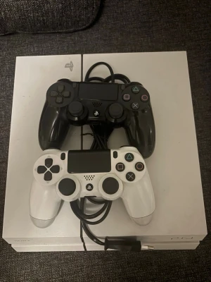 PlayStation 4 med två kontroller - Säljer en vit PlayStation 4 med två DualShock 4-kontroller, en svart och en vit. Konsolen har en stilren design i plast och är perfekt för gaming med vänner. Strömkabel medföljer. Perfekt för dig som vill ha en kraftfull spelkonsol.