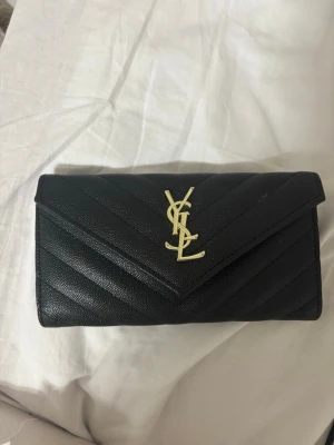Svart YSL plånbok i läder - Snygg svart plånbok från YSL i quiltat läder med guldfärgad logga framtill. Klassisk kuvertform och elegant design som ger en lyxig känsla. Perfekt för dig som vill ha något stilrent och exklusivt i väskan.