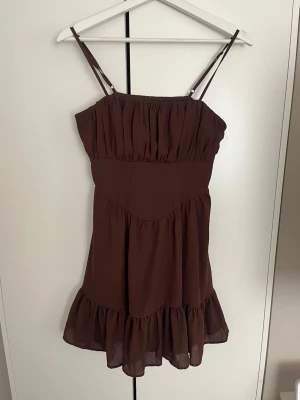 Brun volangklänning med smala axelband - Söt brun kortklänning med smala justerbara axelband och volangkant nedtill. Klänningen har en luftig passform och rynkade detaljer upptill😊Knappt använd då den inte kommer till användning🫶🏼