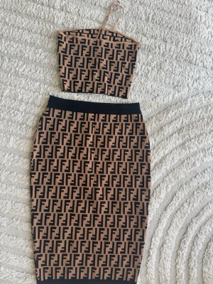 Beige och svart set med FF-mönster - Säljer ett trendigt tvådelat set med crop top och pennkjol i beige och svart. Setet har ett iögonfallande FF-mönster över hela plagget och svarta detaljer i midjan och nederkant. Perfekt för dig som gillar statement-plagg och vill sticka ut.