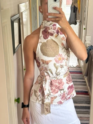 Blommig halterneck topp med knyt - Säljer en snygg halterneck topp med blommigt mönster i rosa, beige och lila toner. Toppen har hög krage, knyt i midjan och är ärmlös. Perfekt för sommarens alla tillfällen och ger en trendig och fräsch look. Jag har sytt Toppen själv och mitt varumärke heter SOWA. Passar xs till M då den är justerbar! 