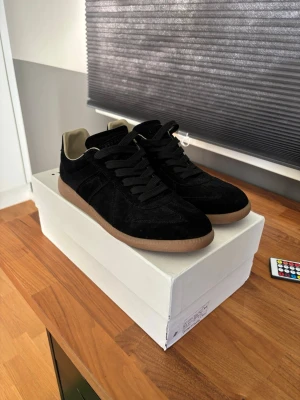 Maison Margiela Gats - Snygga svarta sneakers i mocka med klassisk gummisula och svarta skosnören. Skorna har låg profil och beige innerfoder som ger en stilren kontrast. Perfekta för dig som gillar en clean och tidlös look. Nästintill nya Storlek:44