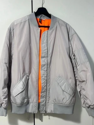 Ljusgrå bomberjacka med orange foder - Snygg ljusgrå bomberjacka med klassisk ribbad krage och muddar. Jackan har ett iögonfallande orange foder, två snedställda fickor framtill och en ärmficka med dragkedja. Tillverkad i ett glansigt syntetmaterial som ger en modern look.