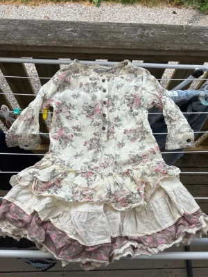 Blommig volangklänning - Supersöt kortklänning i ljusa toner med blommigt mönster och flera lager volanger. Klänningen har knappar framtill, spetsdetaljer och trekvartsärm. Storlek 128cm