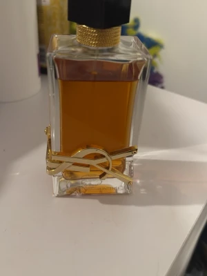 Lyxig parfymflaska med gulddetaljer - Elegant parfymflaska i genomskinligt glas med bärnstensfärgad vätska. Flaskan har en unik geometrisk form och dekoreras med en iögonfallande guldfärgad metallapplikation runt botten. Svart och guld kork ger en exklusiv känsla.
