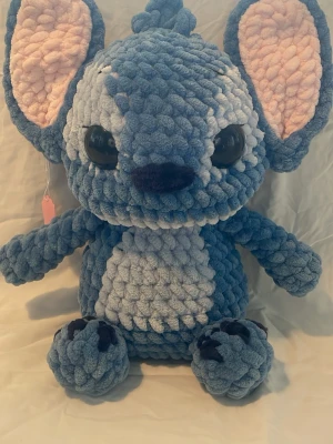 Stor blå virkad Stitch-inspirerad nalle - Säljer en stor, handgjord virkad nalle inspirerad av Stitch. Den är blå med ljusblå mage och ansikte, stora svarta ögon och rosa insida på öronen. Mjuk och fluffig plysch, perfekt som prydnad eller gosedjur. 37 cm hög