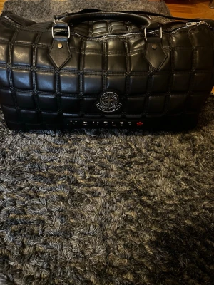 Svart quiltad resväska Moncler - Stone Island - Snygg svart quiltad resväska från Moncler - Stone Island med tydlig logga framtill. Väskan har ett rutmönstrat vadderat yttre, dubbla handtag och dragkedja upptill. Perfekt för weekendresor och har en modern, exklusiv look. Helt oanvänd!