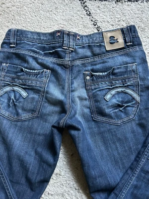 Mörkblå bootcut jeans - Säljer ett par mörkblå bootcut jeans med snygga slitningar och kontrastsömmar. Jeansen har låg midja, fem fickor och detaljerade bakfickor med broderi. Perfekta för en avslappnad och trendig look!! Säljer då de tyvärr är för små för mig💛midje mått är 40cm tvärsöver.