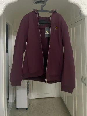 Vinröd hoodie från Lyle & Scott - Säljer en vinröd hoodie från Lyle & Scott med dragkedja framtill och klassisk gul logga på bröstet. Hoodien har huva, två fickor och ribbade muddar. Perfekt för dig som vill ha en stilren och bekväm look.