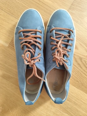 Blå sneakers med bruna skosnören - Snygga sneakers i blå mocka med kontrasterande bruna skosnören och vit sula. Skorna har en klassisk låg design och rund tå. Perfekta för dig som gillar stilrena och avslappnade sneakers med en twist.