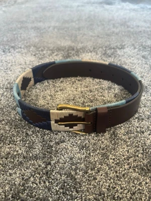 Brunt och blått mönstrat skärp - Snyggt bälte i brunt skinn med detaljer i blått, beige och marinblått mönster. Bältet har en klassisk metallspänne och coola kontrasterande färger som gör det till en unik accessoar till jeans eller byxor.