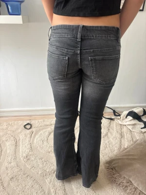 Grå  bootcut jeans - Säljer ett par grå bootcut jeans med slitningar över knäna och råa detaljer. Jeansen har fem fickor och en tvättad look som ger en cool vibe. Perfekta för dig som gillar en avslappnad och trendig stil.