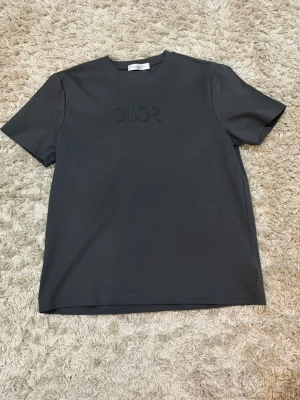 Svart t-shirt från Dior med logga - Snygg svart t-shirt från Dior med diskret logga i samma färg på bröstet. Klassisk rund halsringning och korta ärmar. Tillverkad i mjuk bomull för en bekväm känsla och stilren look.