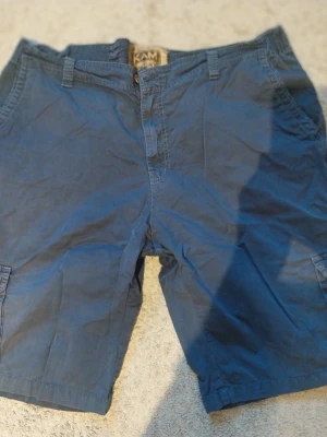 Blå cargoshorts från KAM - Säljer ett par blå cargoshorts från KAM med klassisk passform. Shortsen har flera fickor, bälteshällor och dragkedja framtill. Perfekta för sommardagar och avslappnade tillfällen.