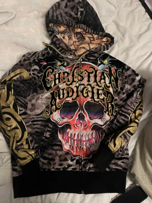 Christian Audigier hoodie med dödskalle - Säljer en riktigt fet hoodie från Christian Audigier med stort dödskalletryck i rött och lila på framsidan, leopardmönster i grått och svart samt guldiga detaljer på ärmarna. Huvan har ett unikt mönster på insidan och dragkedja framtill. Perfekt för dig som gillar streetwear.