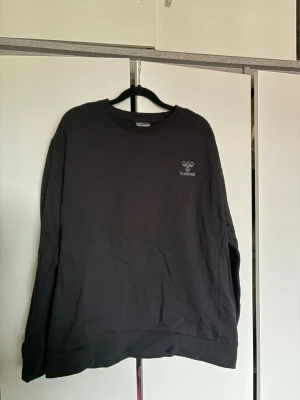 Svart sweatshirt från Hummel - En stilren svart sweatshirt från Hummel med rund halsringning och diskret logga på bröstet. Tröjan har en klassisk passform och ribbade muddar vid ärmslut och nederkant. Perfekt för en avslappnad och sportig look.