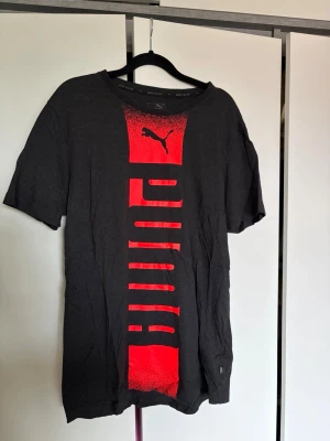 Svart t-shirt från Puma med röd logga - Svart t-shirt från Puma med en stor röd vertikal logga och Puma-tryck framtill. 
