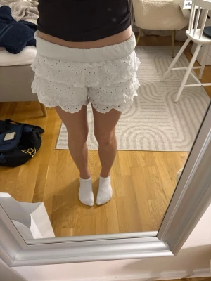 Vita volangshorts med broderi - Supersöta vita shorts med flera lager volanger och broderade detaljer. De har elastisk midja och är perfekta för varma dagar. Stilen är feminin och luftig, med fina spetskanter och ett romantiskt uttryck.
