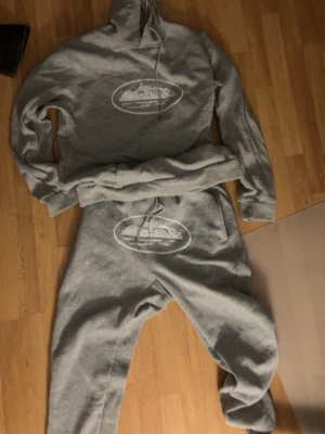 Grått loungewear-set med tryck - Mysigt grått loungewear-set med hoodie och byxor. Båda delarna har ett vitt tryck med motiv av ett fartyg i en oval ram. Hoodien har dragsko i huvan och byxorna har resår i midjan samt dragsko. Perfekt för chill dagar hemma.