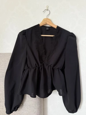 Svart blus med spetsdetaljer från SHEIN - Svart blus från SHEIN med djup v-ringning och spetsdetaljer längs urringningen. Blusen har långärmade puffärmar och ett lätt, flödande tyg som ger en luftig känsla. Perfekt för dig som gillar en chic och trendig look.