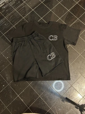 Svart set med CB-tryck - Säljer ett svart set med t-shirt och shorts, båda med vitt CB-tryck på bröstet och benet. Setet har en enkel och avslappnad design med elastisk midja på shortsen. Perfekt för en chill och stilren look.