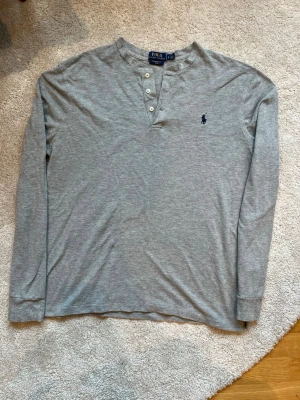 Grå Polo Ralph Lauren tröja  - Snygg grå långärmad tröja från Polo Ralph Lauren med tre knappar vid halsen med mörkblå broderade loggan på bröstet. Passar som storleken M. Perfekt för en avslappnad och stilren look.