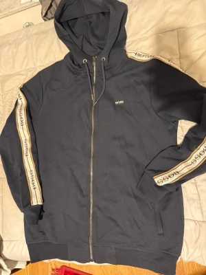 Marinblå hoodie från BOSS med dragkedja - Snygg marinblå hoodie från BOSS med dragkedja framtill och huva med dragsko. Ärmarna har vita och guldiga band med BOSS-logga längs sidorna. Tillverkad i mjukt bomullsmaterial och har fickor på sidorna. Perfekt för en chill och stilren look.