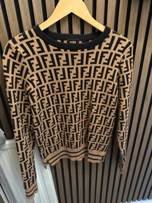 Beige och svart stickad Fendi-tröja - Stickad tröja från Fendi i beige och svart med det ikoniska FF-mönstret över hela plagget. Rund halsringning och ribbade muddar vid ärmslut och nederkant. Perfekt för dig som vill ha en trendig och lyxig look.