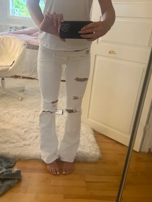 Vita slitna low waist jeans - Säljer ett par vita bootcut jeans med slitningar över knäna och låren. Jeansen har låg midja och detaljerade bakfickor med broderi. Perfekta för dig som gillar en avslappnad och trendig stil.