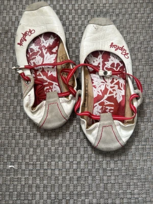 Vita Replay ballerinaskor med röd detalj - Säljer ett par vita ballerinaskor från Replay med röda detaljer och logga. Skorna har röd insida med blommönster, röda elastiska band och grå mockadetaljer på tån. Perfekta för dig som gillar en sportig och unik stil.