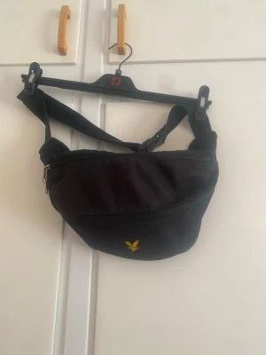 Svart Lyle & Scott axelväska  - Snygg svart axelväska i tyg med justerbart band och dragkedja. Väskan har en gul broderad logga framtill och en enkel, sportig design. Perfekt för att bära det viktigaste och passar till många olika stilar.