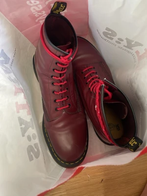 Vinröda Dr. Martens boots - Säljer ett par klassiska vinröda Dr. Martens boots i skinn med svarta och gula detaljer samt röda skosnören. Bootsens ikoniska gula söm syns runt sulan och de har en högre modell med dragflik bak. Perfekta för dig som vill sticka ut med din stil.